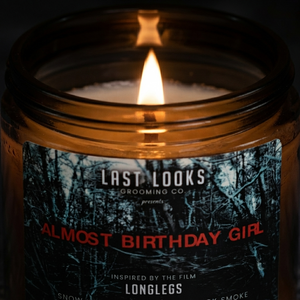 A lit amber glass jar candle with a white wick and a dark forest-themed label reading 'LAST LOOKS GROOMING CO presents ALMOST BIRTHDAY GIRL' in red text.