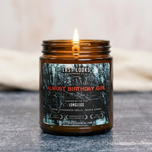 A lit amber glass jar candle with a black and red label reading 'ALMOST BIRTHDAY GIRL' by Last Looks Grooming Co, set on a gray textured surface with a soft beige background.