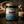 A glass jar candle with a gold metal lid sits on a rustic wooden surface, surrounded by coiled rope and driftwood. The candle has a label featuring the brand 'Last Looks Grooming Co.
