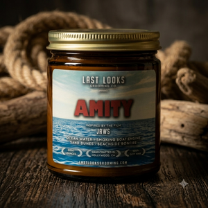 A glass jar candle with a gold metal lid sits on a rustic wooden surface, surrounded by coiled rope and driftwood. The candle has a label featuring the brand 'Last Looks Grooming Co.