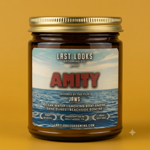A single amber glass jar candle with a gold metal lid and a custom ocean-themed label reading 'LAST LOOKS GROOMING CO. AMITY' and 'INSPIRED BY THE FILM JAWS,' centered on a warm mustard-yellow background.