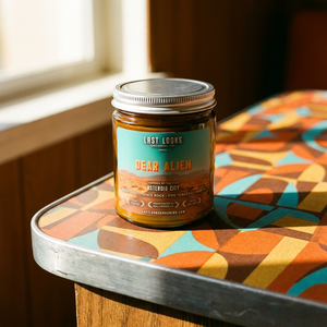 A glass jar candle with a silver metal lid and a teal label reading 'DEAR ALIEN' sits on a patterned table in a warm indoor setting near a window.