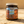 A glass jar candle with a silver metal lid, featuring a colorful label with an illustration of Mediterranean buildings and the text 'LAST LOOKS GROOMING CO. ELIO'.