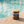A glass jar candle labeled 'Last Looks Hand Grenade' with a gold lid and amber-brown wax, placed on a stone surface with a blurred turquoise pool in the background.