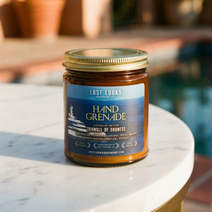 A glass jar candle with a gold metal lid sits on a white marble table outdoors. The candle has a blue and white label reading 'LAST LOOKS GENEVAISE HAND GRENADE' with a boat graphic.