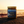 A jar candle labeled 'Last Looks Hand Grenade' sits on a dark textured surface with a blurred beach sunset background.