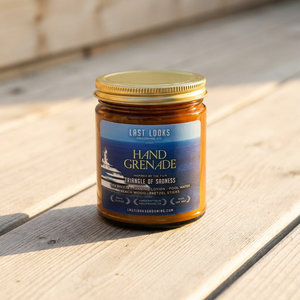 A glass jar candle with a gold lid and a blue label reading 'Last Looks Hand Grenade Triangle of Sronness' sits on a light wooden deck outdoors.