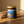 A glass jar candle with a gold lid and a blue label reading Last Looks Hand Grenade, placed on a wooden surface with cinnamon sticks beside it in warm sunlight.