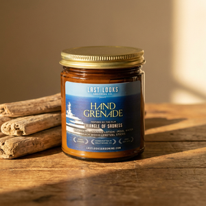 A glass jar candle with a gold lid and a blue label reading Last Looks Hand Grenade, placed on a wooden surface with cinnamon sticks beside it in warm sunlight.
