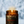 An amber glass jar candle with a lit wick, featuring a dark forest-patterned label with red and white text, set against a blurred snowy outdoor background.