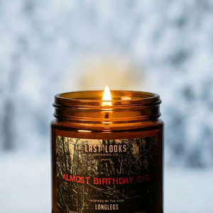 An amber glass jar candle with a lit wick, featuring a dark forest-patterned label with red and white text, set against a blurred snowy outdoor background.