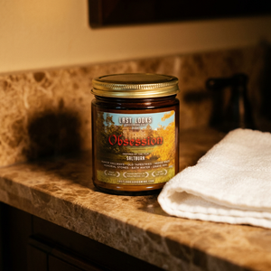 A Saltburn candle in a dark amber glass jar with a gold lid sits on a brown marble countertop next to a folded white towel. The label reads 'Last Looks Candle Co.