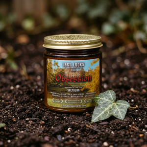A glass jar of Last Looks Saltburn candle with a gold lid sits on dark soil next to a green ivy leaf, with a blurred natural background.
